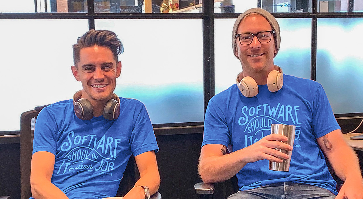 Two young men wearing matching blue shirts with the word "software" on them, sitting in chairs, they have headphones around their necks. They are smiling.